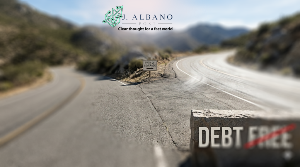 A photorealistic view of a winding mountain road splitting into two paths, with a large stone in the foreground featuring the words "Debt Free" where the word "Free" is crossed out with a red line.